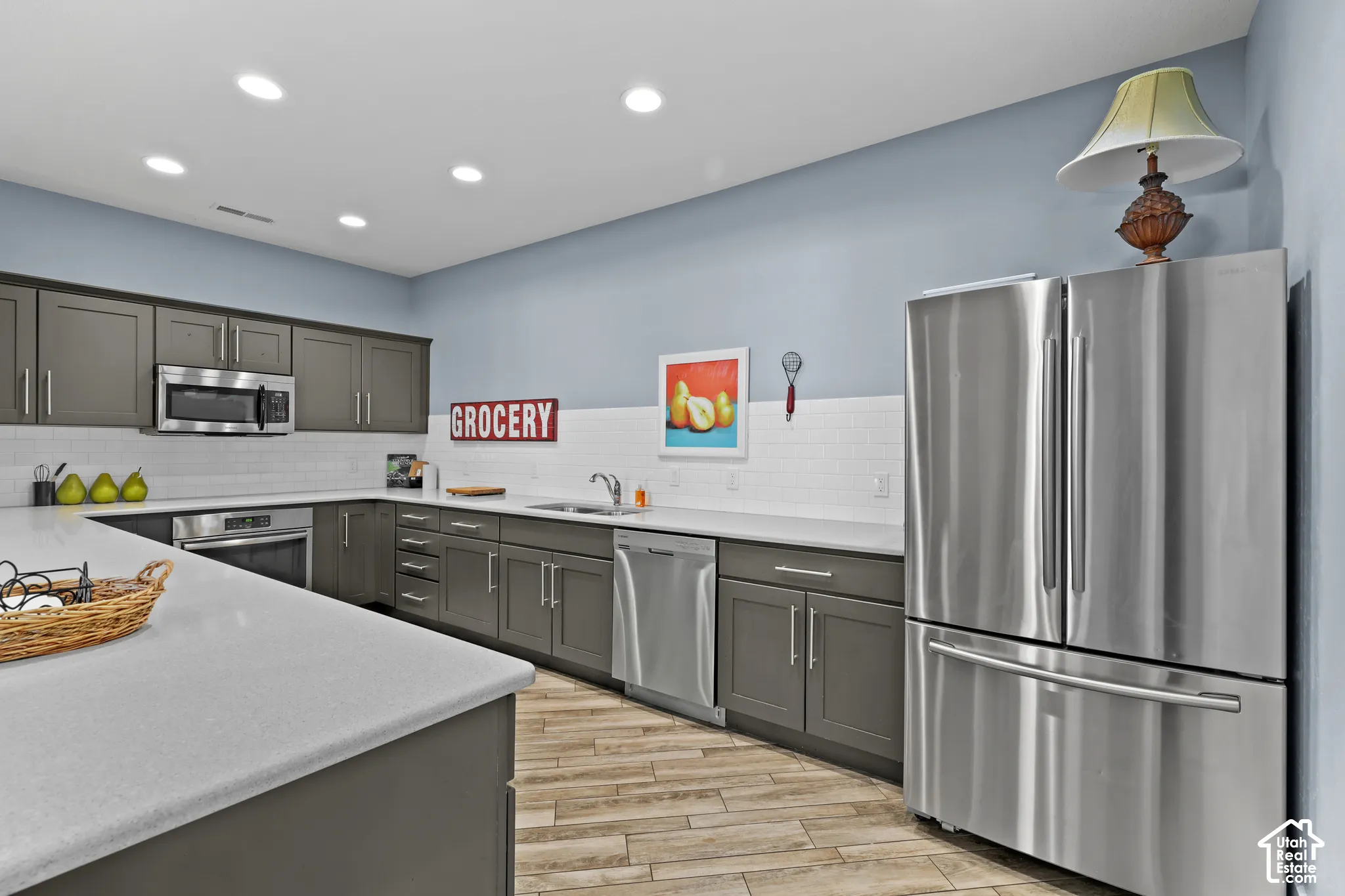 Kitchen featuring dark-toned cabinetry, light-colored countertops, stainless steel appliances, a white subway tile backsplash, and wood-finish flooring