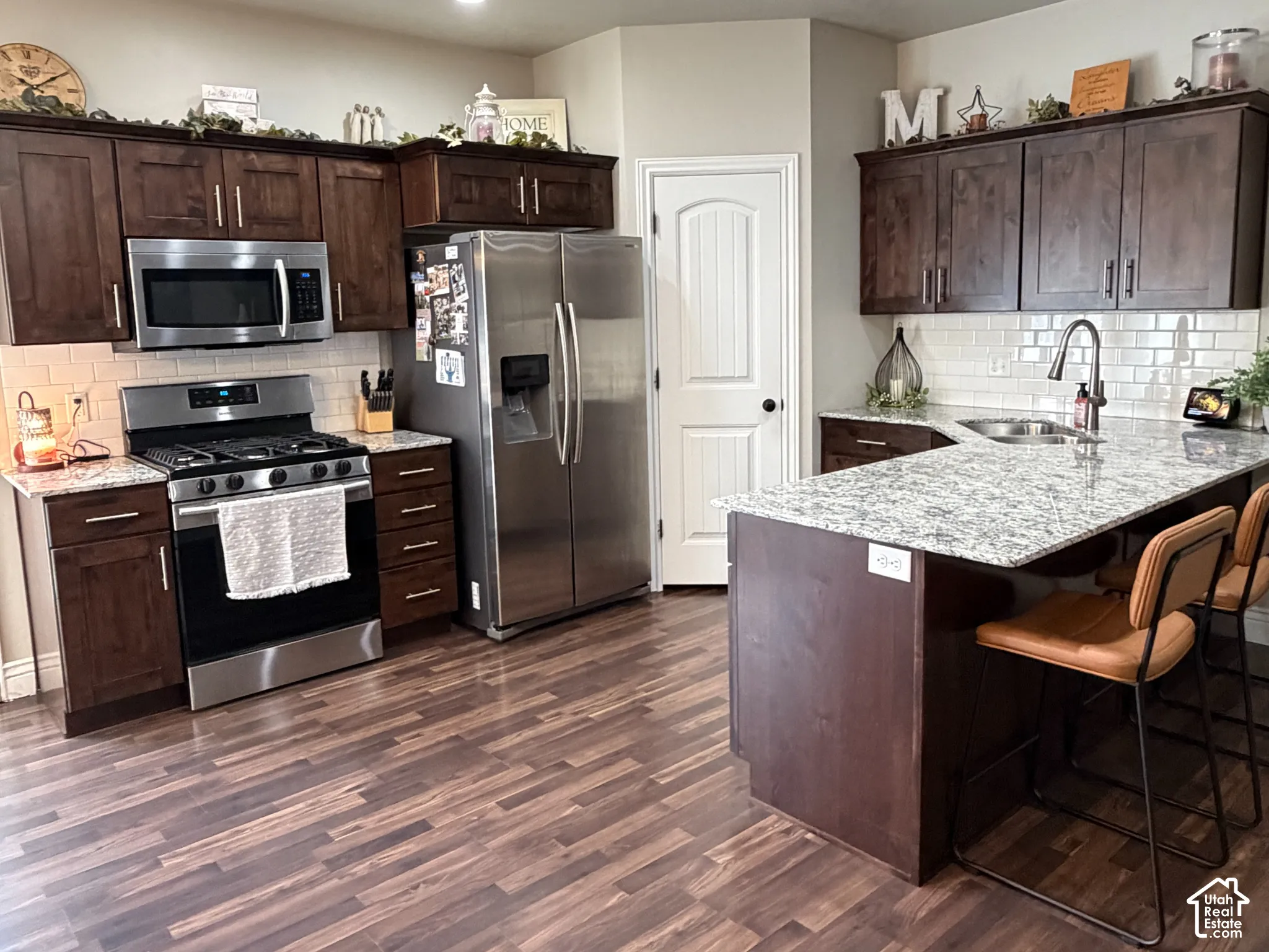 Kitchen featuring dark wood cabinetry, stainless steel appliances, light-toned stone countertops, subway tile backsplash, and wood-finish flooring