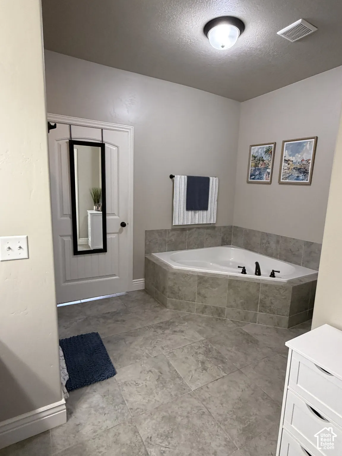Corner soaking tub featuring a tile surround, large-format tile flooring, and a ceiling-mounted light fixture
