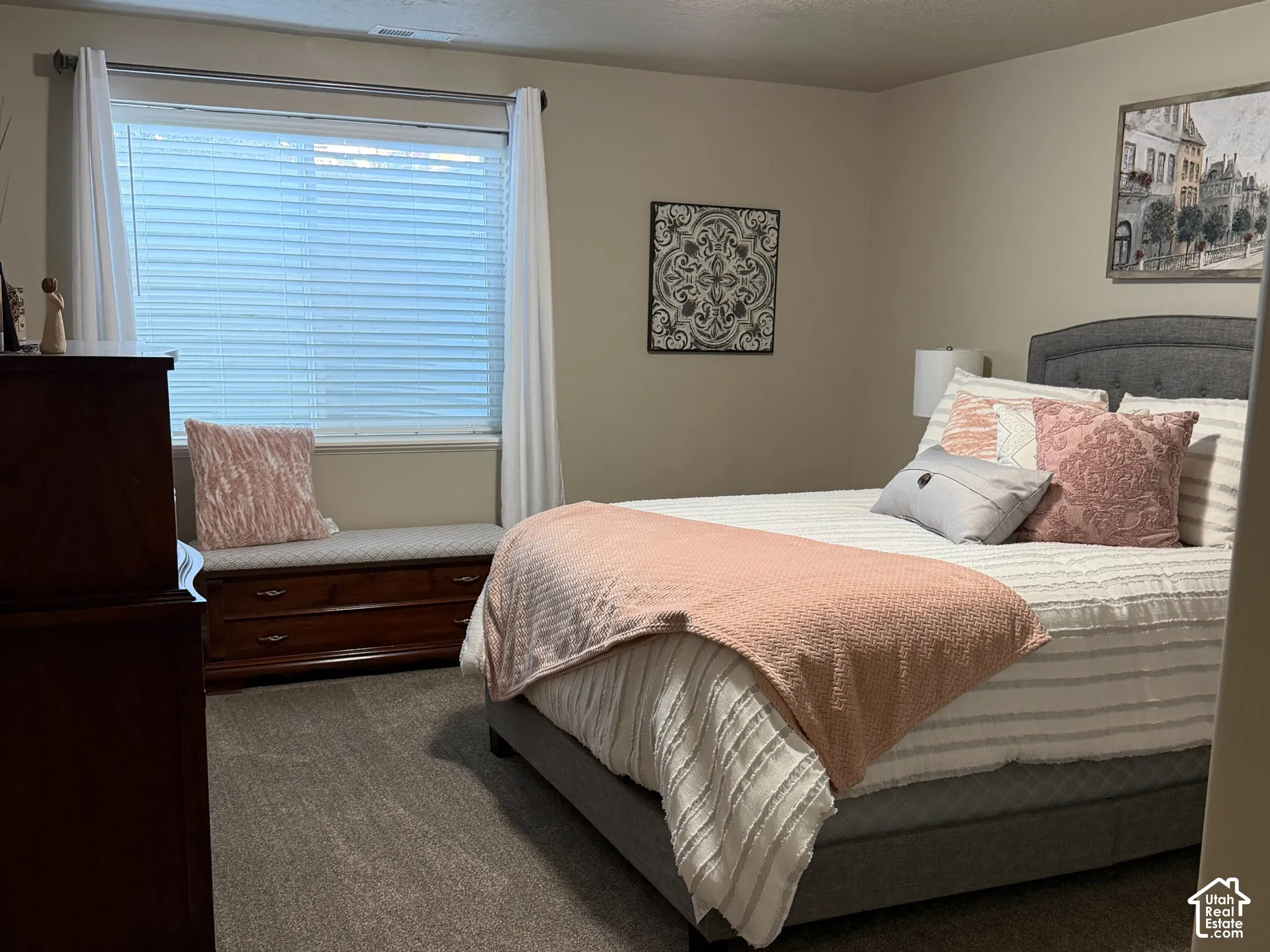 Neutral-toned bedroom featuring wall-to-wall carpeting, a large window with horizontal blinds, and a built-in wooden storage bench beneath the window