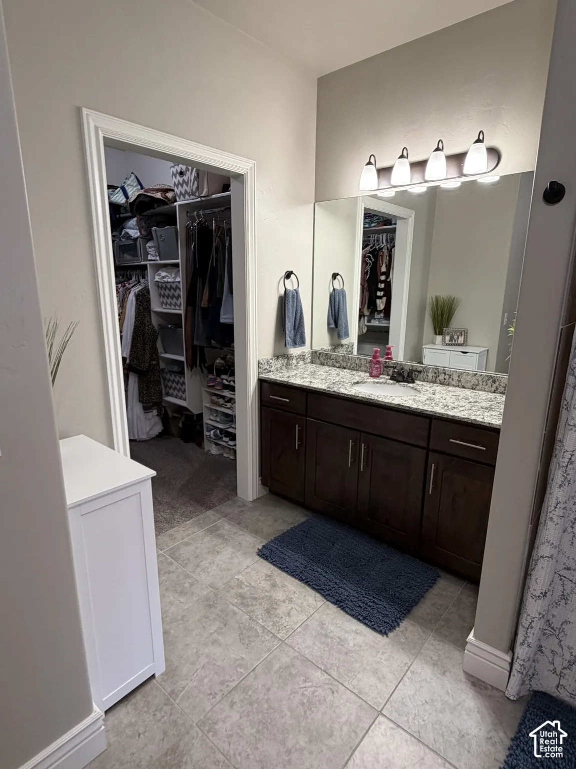 Bathroom featuring a vanity with stone-patterned countertop, dark wood-finish cabinetry, a large frameless mirror, and four-bulb overhead lighting, with entry to a walk-in closet featuring built-in shelving