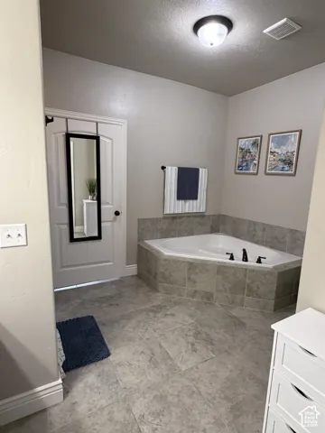 Corner soaking tub featuring a tile surround, large-format tile flooring, and a ceiling-mounted light fixture
