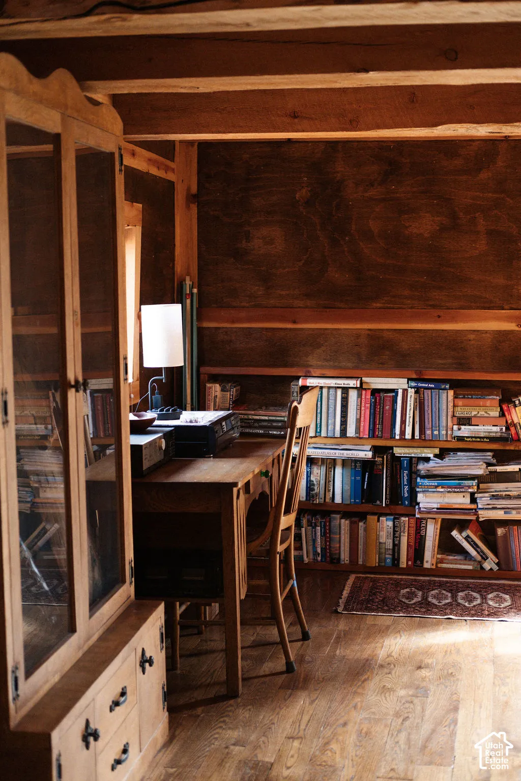 Exposed wood ceiling beams, dark stained wood paneling, built-in open bookshelves, wood-finish flooring, and a floor-to-ceiling built-in wood cabinet with glass door insets