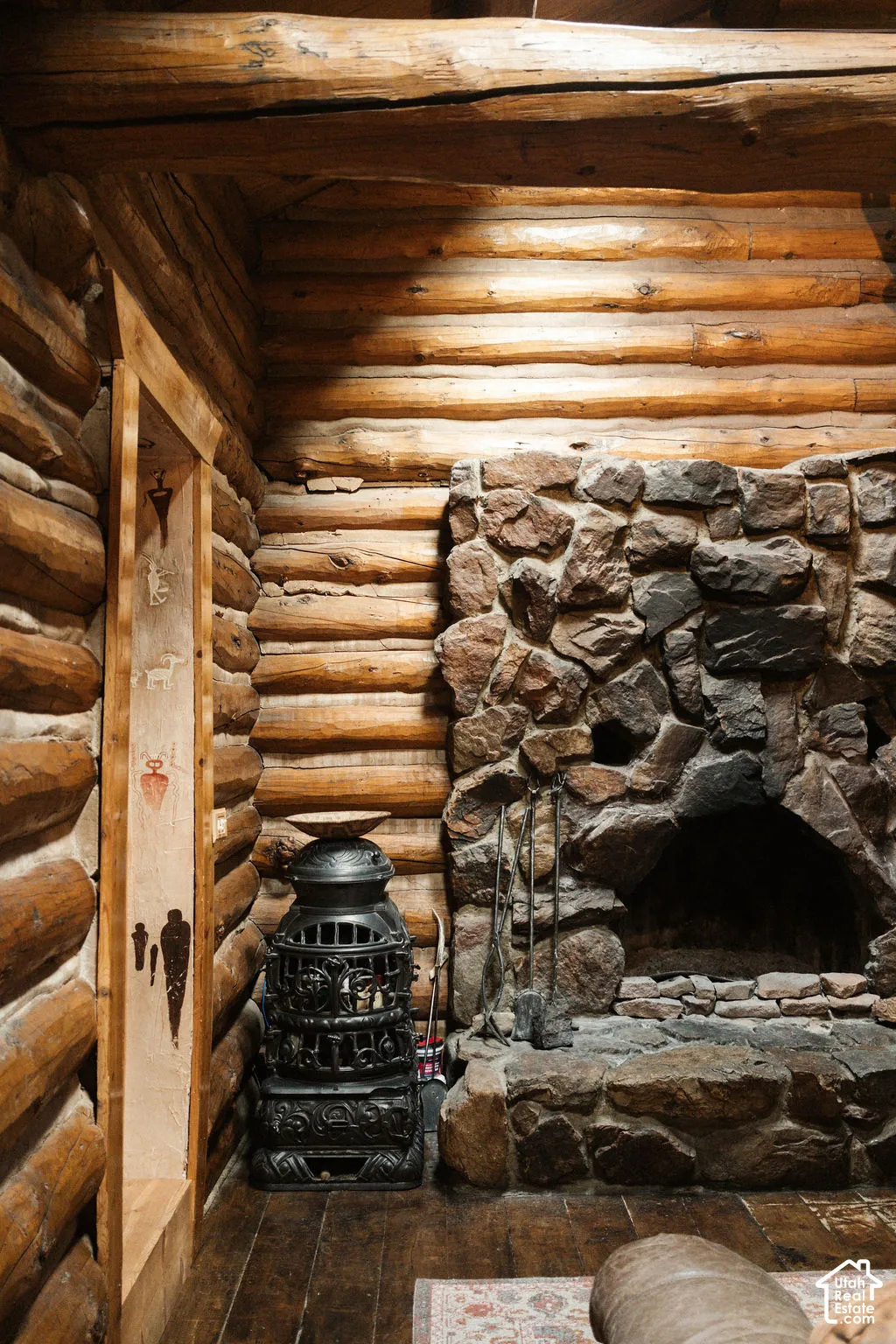 Interior featuring stacked log walls, an exposed timber ceiling beam, a floor-to-ceiling stone fireplace with a raised hearth, and wood-finish flooring