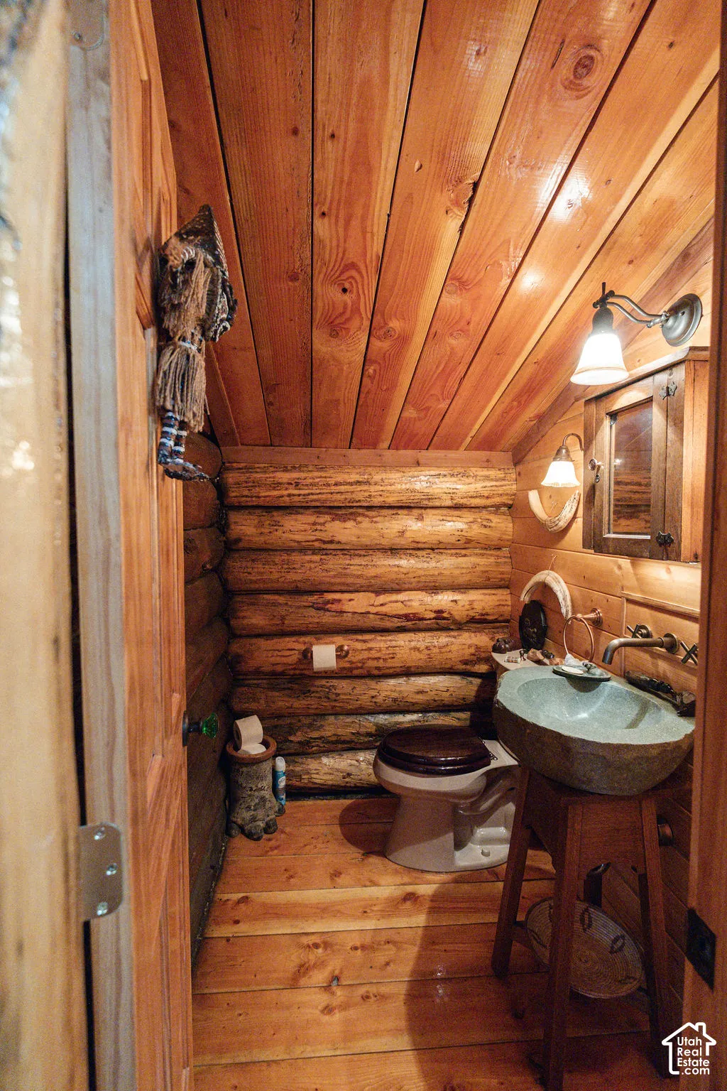 Full-log wall construction, wood-plank ceiling, wood-finish flooring, stone-basin sink on a wooden pedestal, and rustic wall-mounted lighting