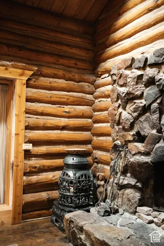 Rustic log walls featuring exposed timber beams, full-height stone fireplace surround with a raised stone hearth, and a decorative cast-iron stove