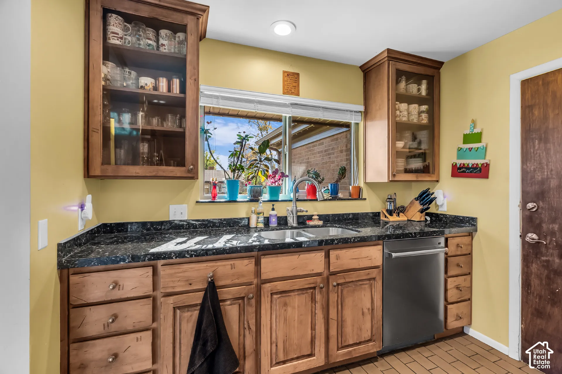 Kitchen area featuring wood cabinetry with glass-front upper displays, dark stone countertops, stainless steel dual-basin sink, integrated stainless steel dishwasher, and ceramic tile flooring