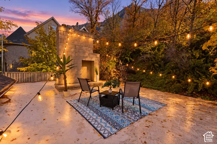 Outdoor stone patio featuring a built-in masonry fireplace, string lighting, and mature hillside landscaping