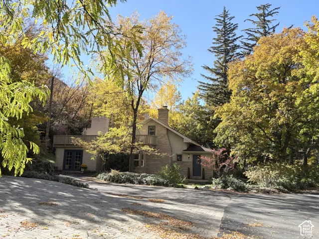 Multi-level exterior featuring a brick facade, integrated chimney, wood-railing balcony, and paved driveway, set among mature trees