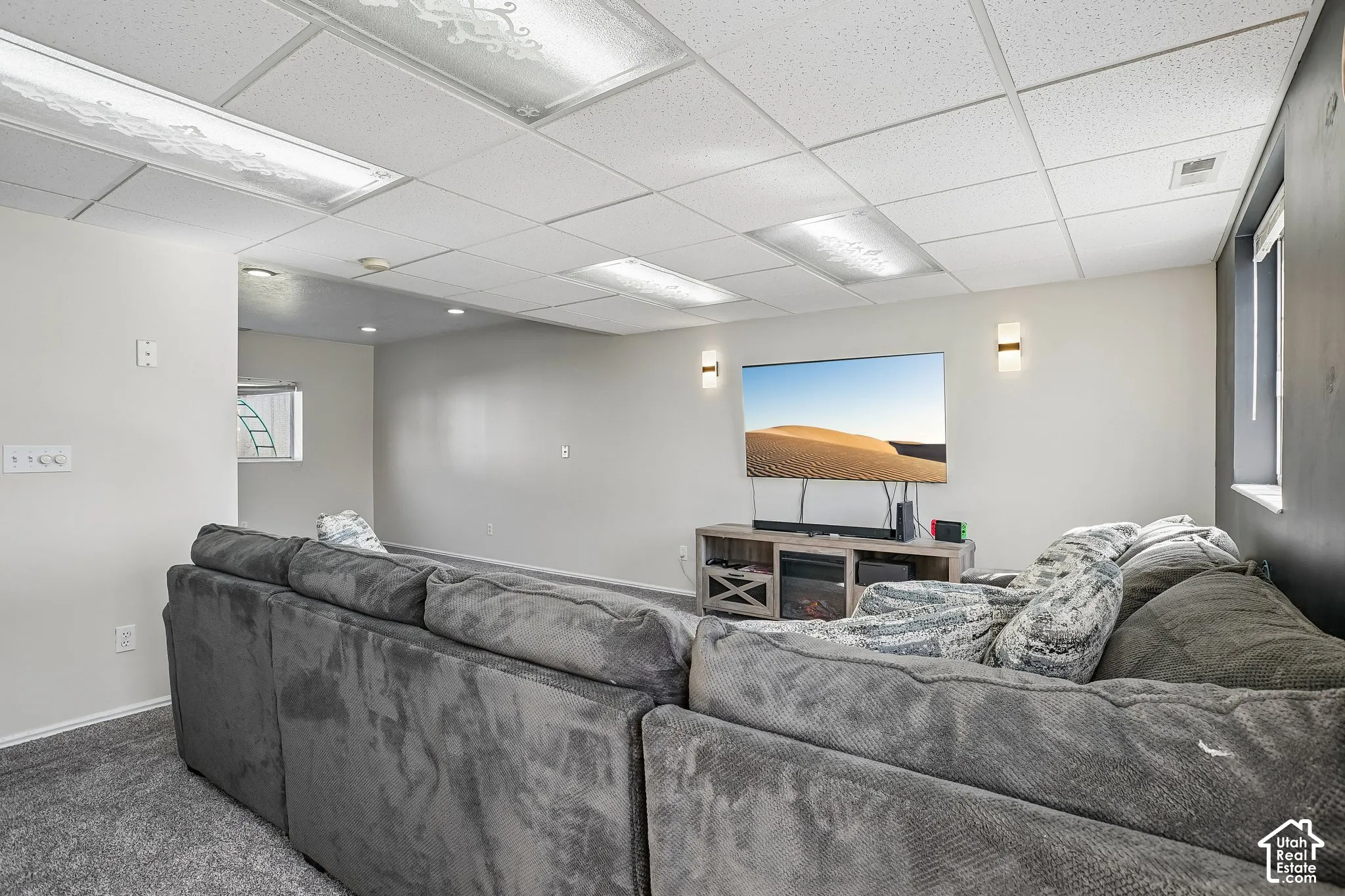 Neutral-toned interior space featuring recessed drop-ceiling panels, flush-mount fluorescent lighting, wall-mounted sconces, and carpeting