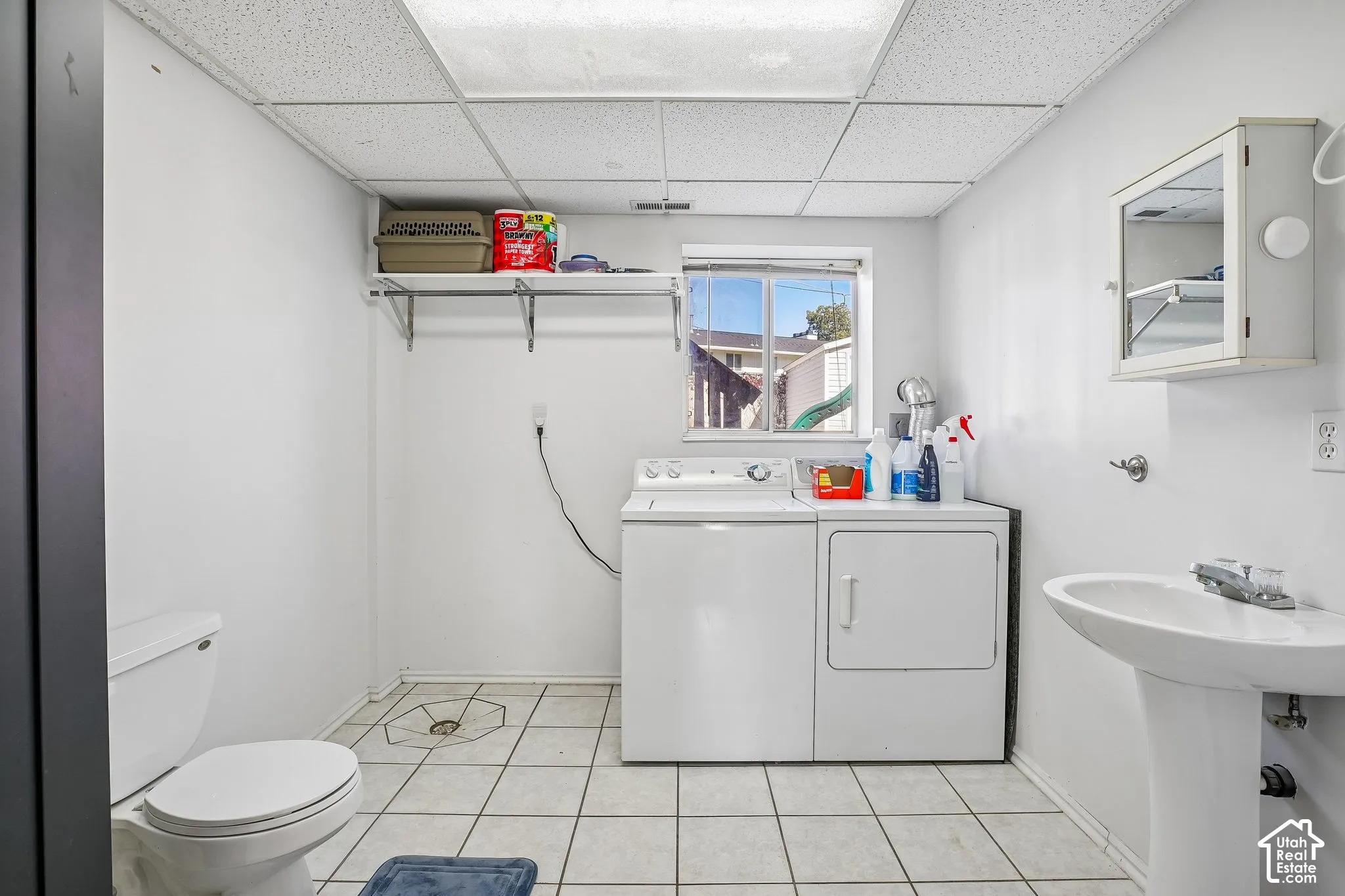 Laundry area featuring ceramic tile flooring, a pedestal sink, wall-mounted storage shelving, and a drop ceiling