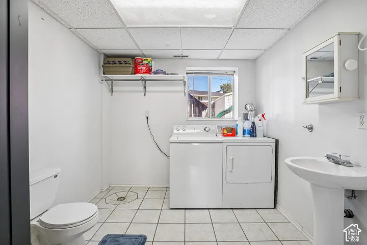 Laundry area featuring ceramic tile flooring, a pedestal sink, wall-mounted storage shelving, and a drop ceiling