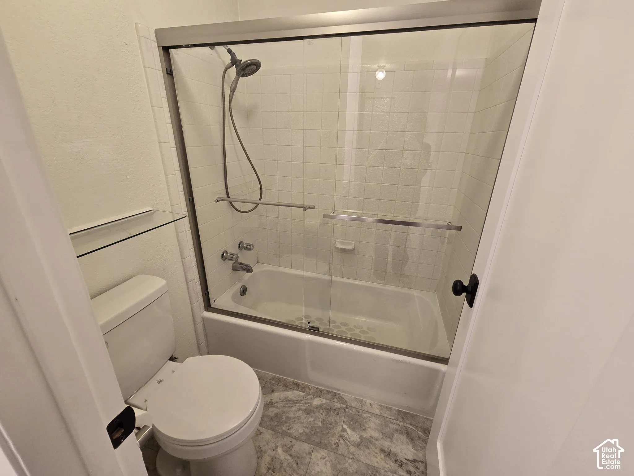 Bathtub and shower combination with floor-to-ceiling tile surround, glass sliding enclosure, adjustable handheld shower head, and stone-finish tile flooring
