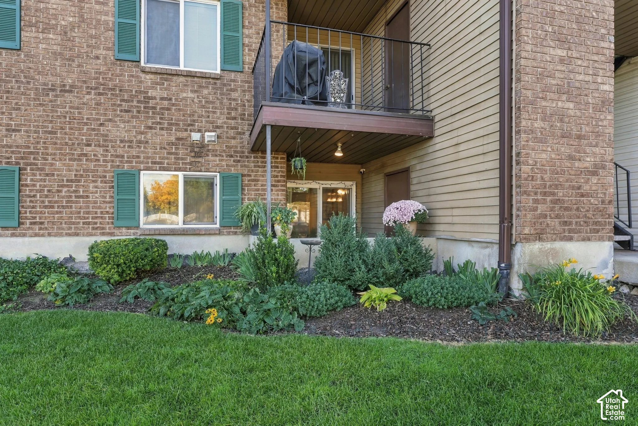 Brick and horizontal siding exterior featuring green shutters, an elevated balcony with metal railings, and a landscaped garden bed