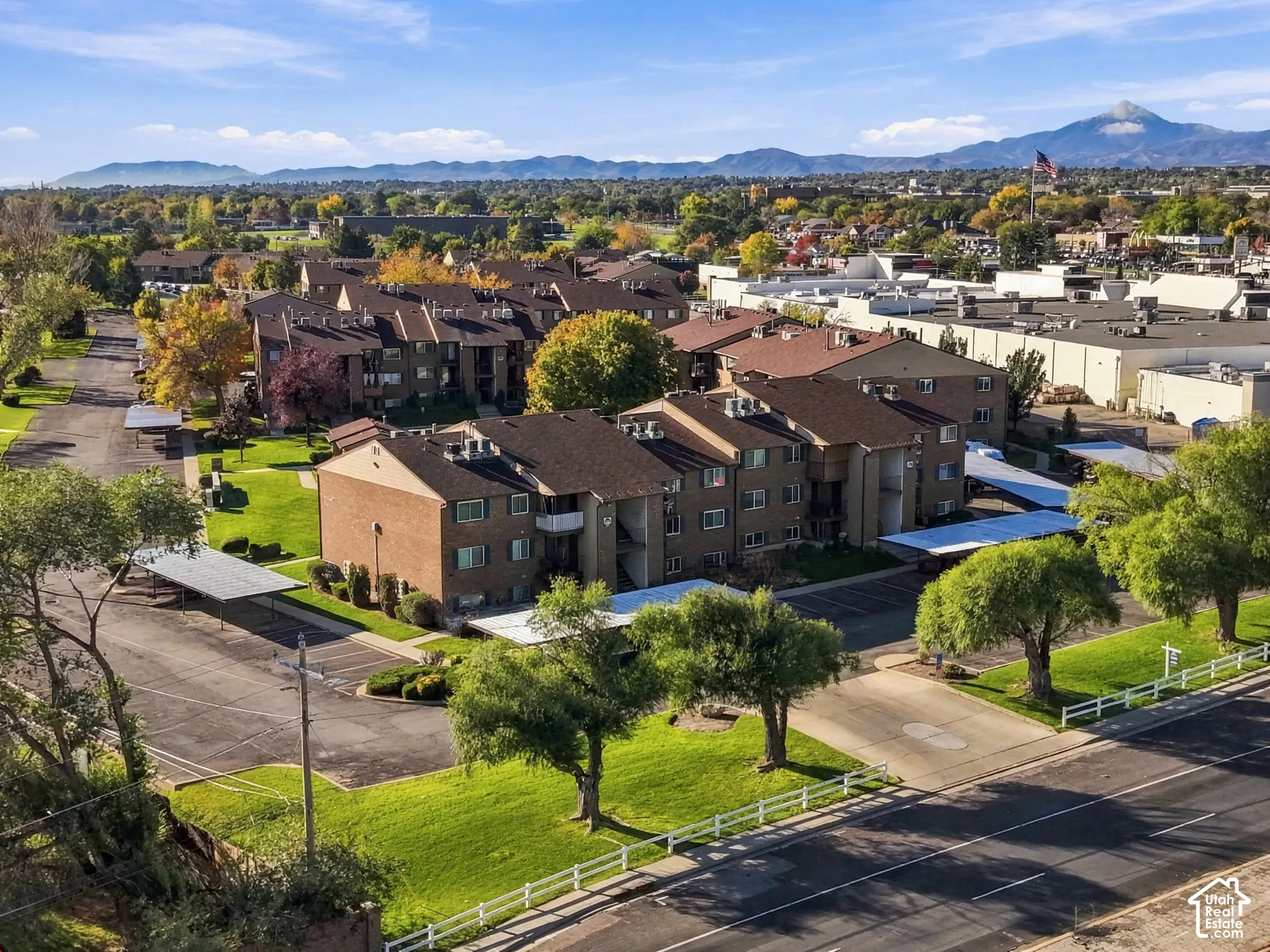 Multi-story brick building exterior featuring covered parking structures, mature trees, manicured landscaping, and mountain backdrop