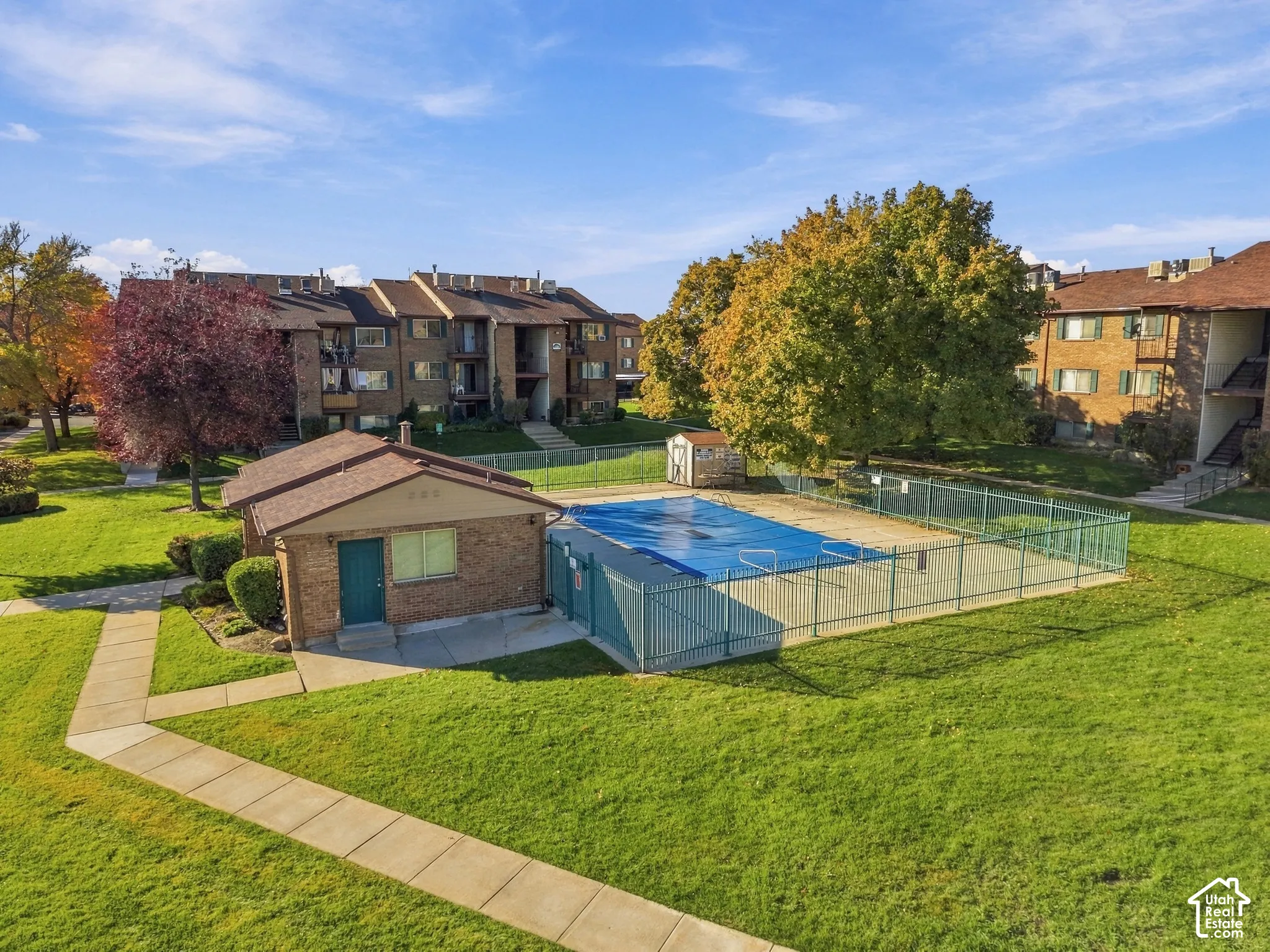 Fenced swimming pool with a covered utility building, situated on expansive grounds featuring manicured lawns, paved walkways, and mature trees