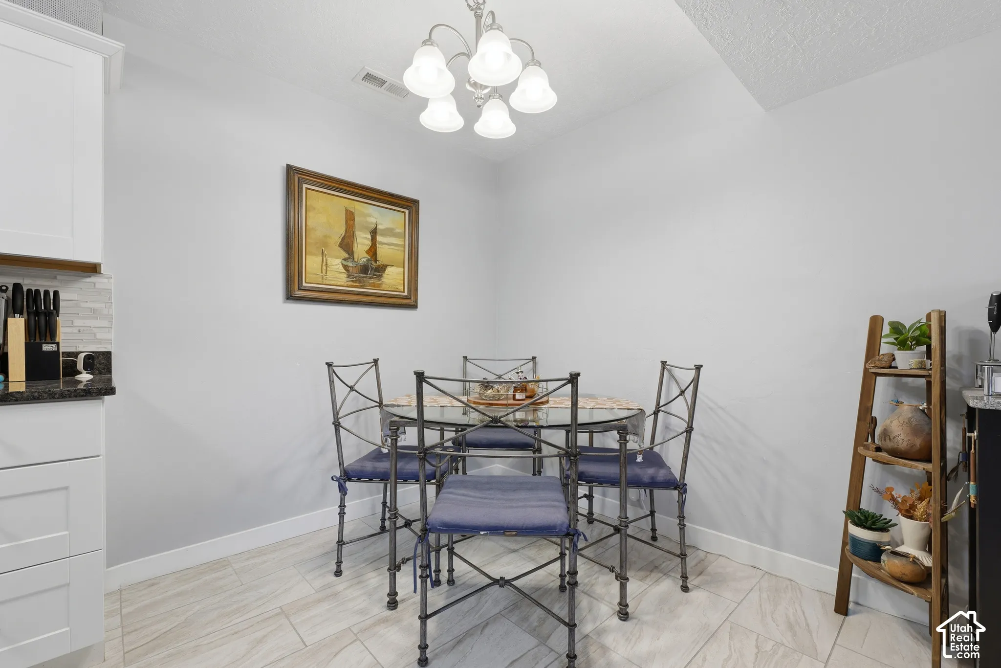 Dining area featuring a multi-bulb chandelier, light-toned tile flooring, and white shaker-style cabinetry with dark countertops