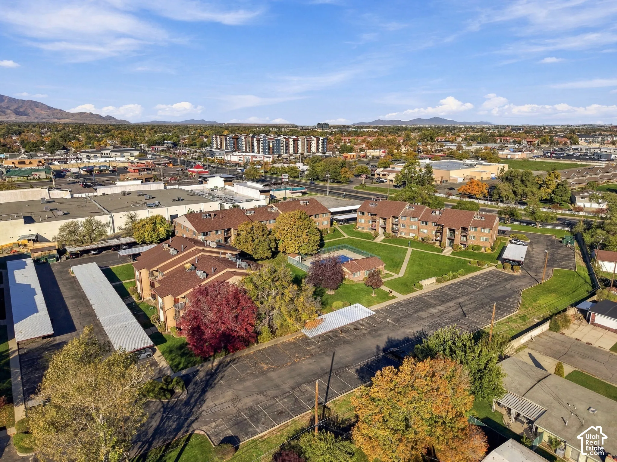 Multi-family residential complex featuring mid-rise brick buildings, paved surface parking lots, covered parking structures, a central communal pool, and mature landscaping