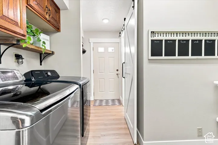 Laundry area featuring wood-finish flooring, white sliding barn door, upper cabinetry with natural wood finish, wall-mounted shelving, and paneled exterior door