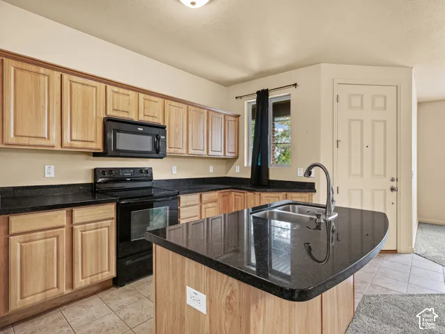 Kitchen featuring light wood cabinetry, dark stone countertops, central island with an integrated dual-basin sink, and black built-in appliances