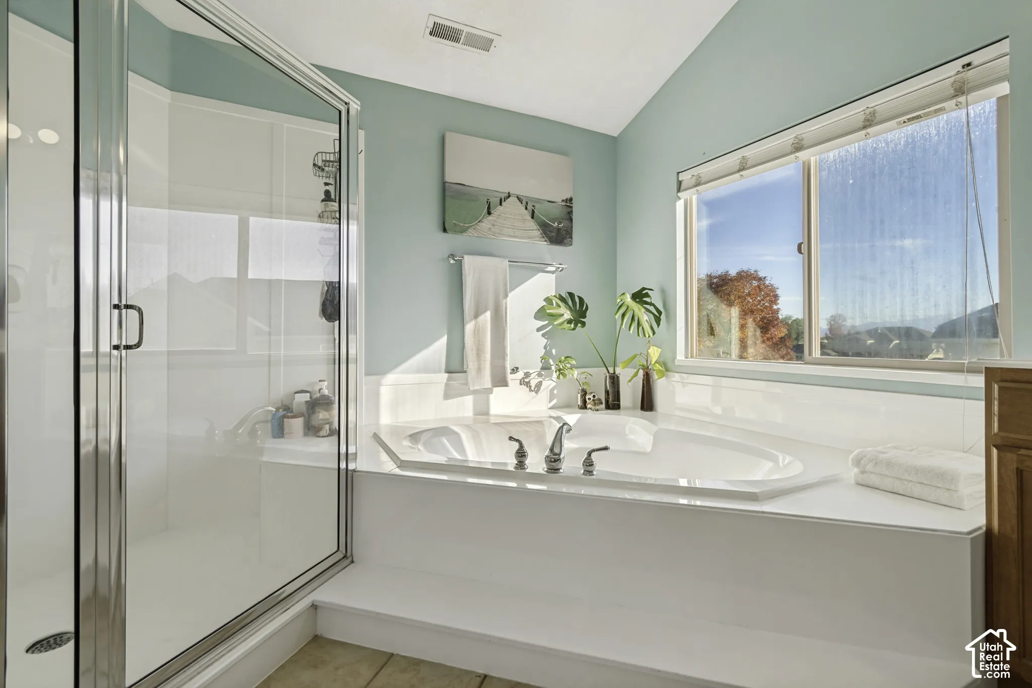Corner soaking tub with a white tile surround, glass-enclosed shower, and large horizontal window