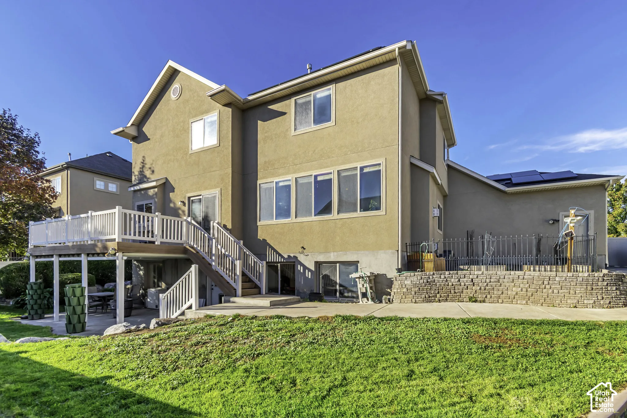 Exterior elevation featuring a raised deck with white railings, a concrete patio, stone retaining wall, and roof-mounted solar panels