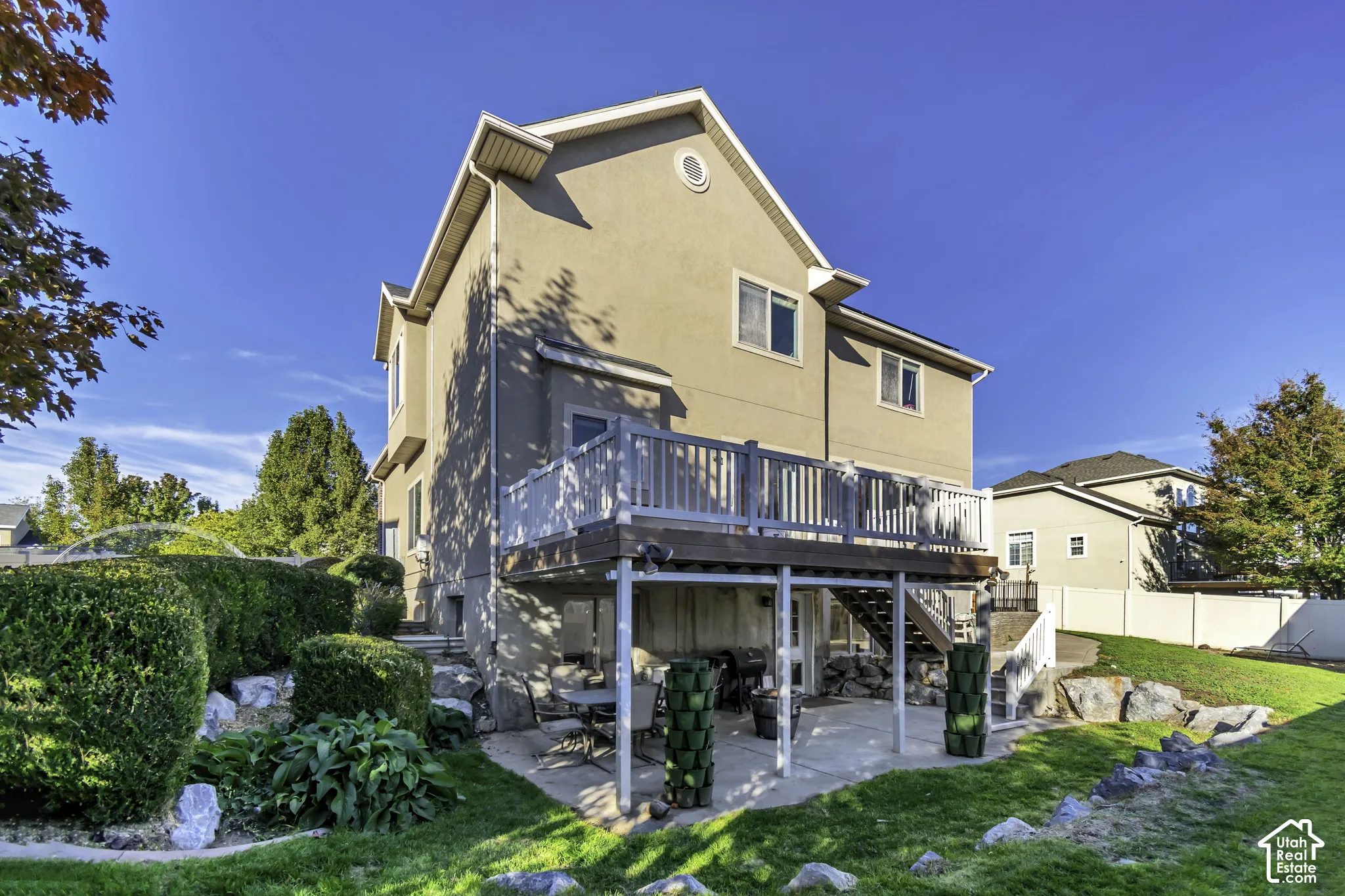 Stucco exterior featuring an elevated deck with white balustrades, a concrete ground-level patio, and natural stone landscaping elements