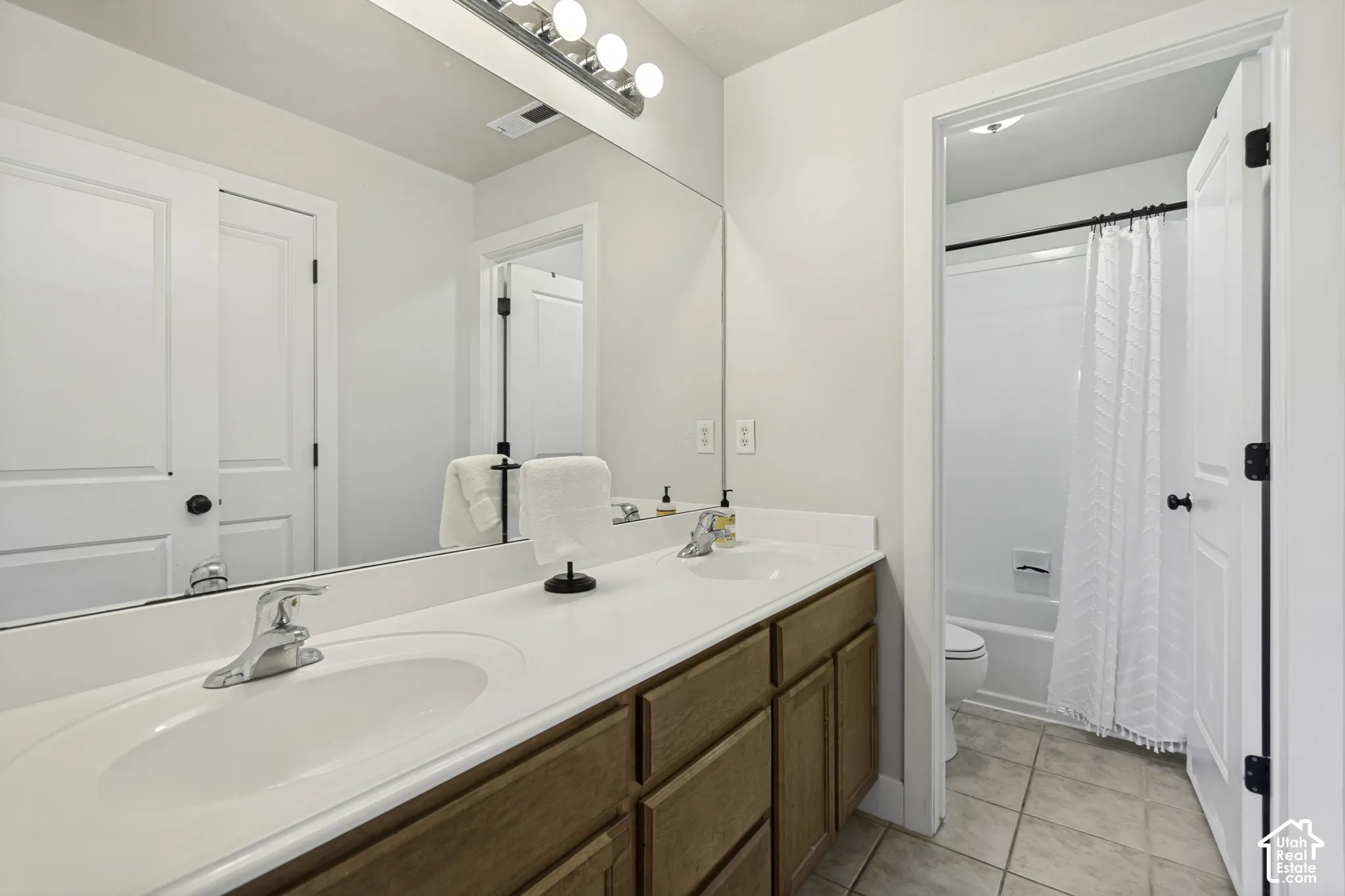 Dual-sink vanity with wood-finish cabinetry, solid-surface countertop, full-width vanity mirror, tile flooring, and a combined tub and shower enclosure