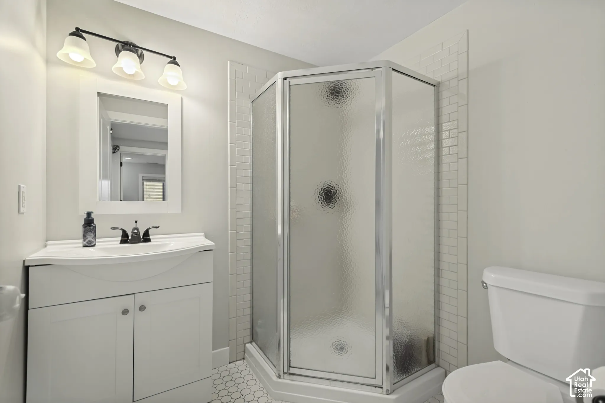 Bathroom featuring a corner shower with textured glass enclosure, white subway tile surround, hexagonal floor tiling, white vanity with integrated sink, and three-light wall-mounted fixture
