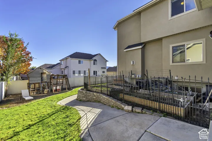 Rear yard featuring a manicured lawn, tiered stone retaining walls, metal fencing, and concrete patio surfacing