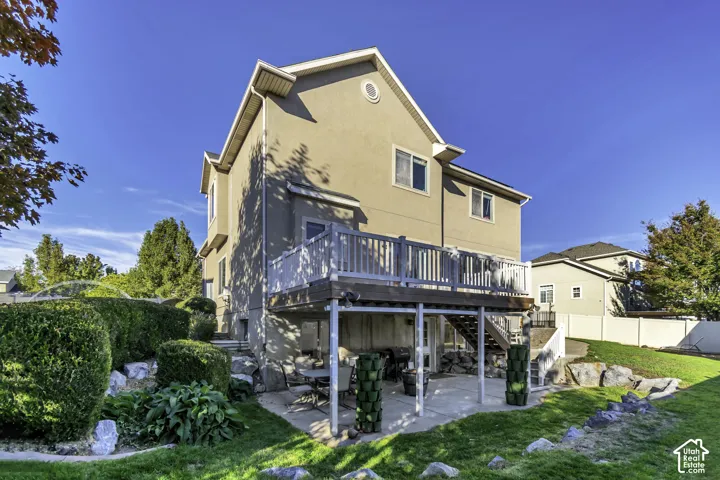 Stucco exterior featuring an elevated deck with white balustrades, a concrete ground-level patio, and natural stone landscaping elements