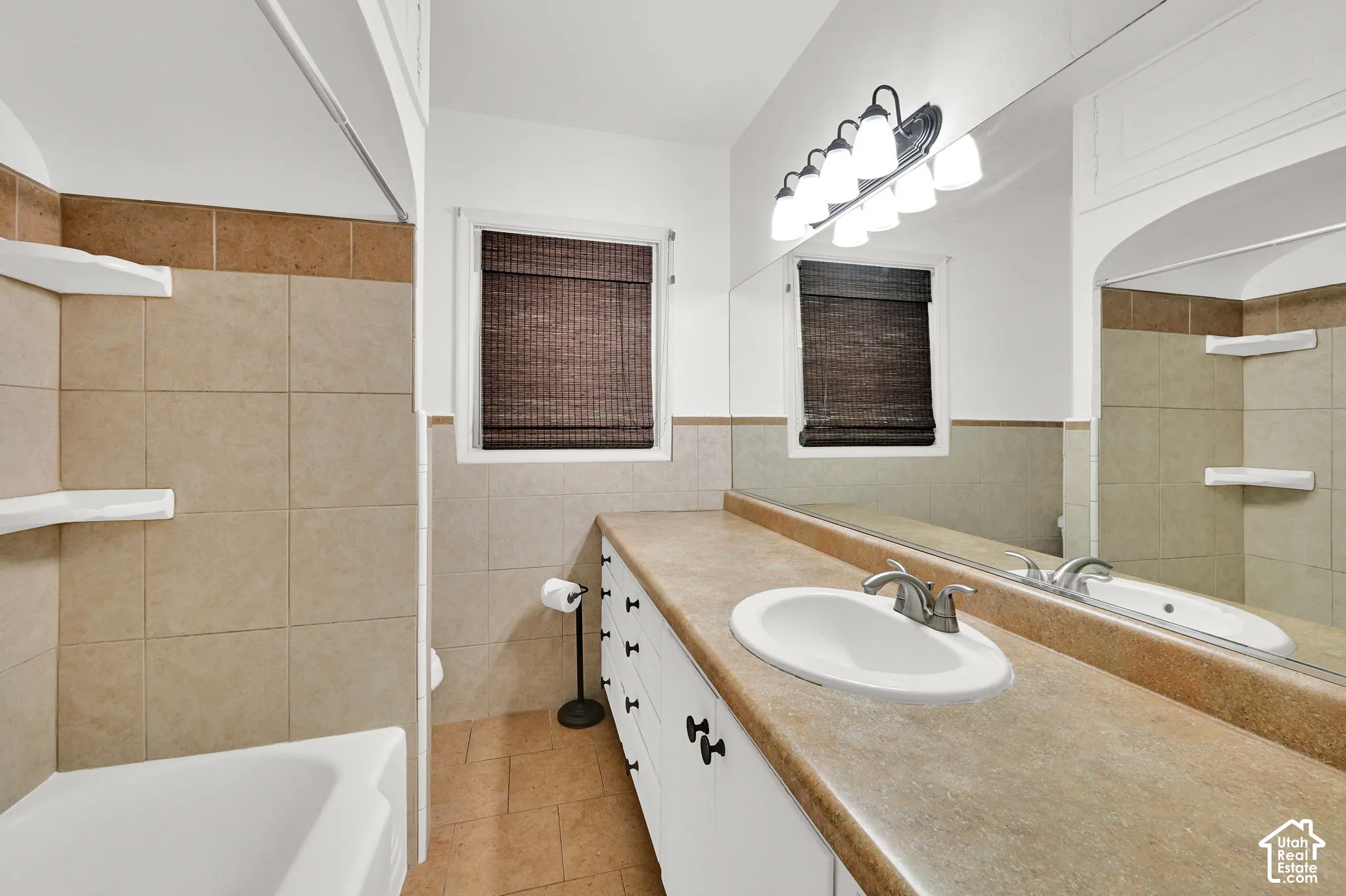 Bathroom featuring a long vanity with white cabinetry, a neutral solid-surface countertop, an integrated oval sink, full-width wall mirror, and ceramic tile surround in the shower area