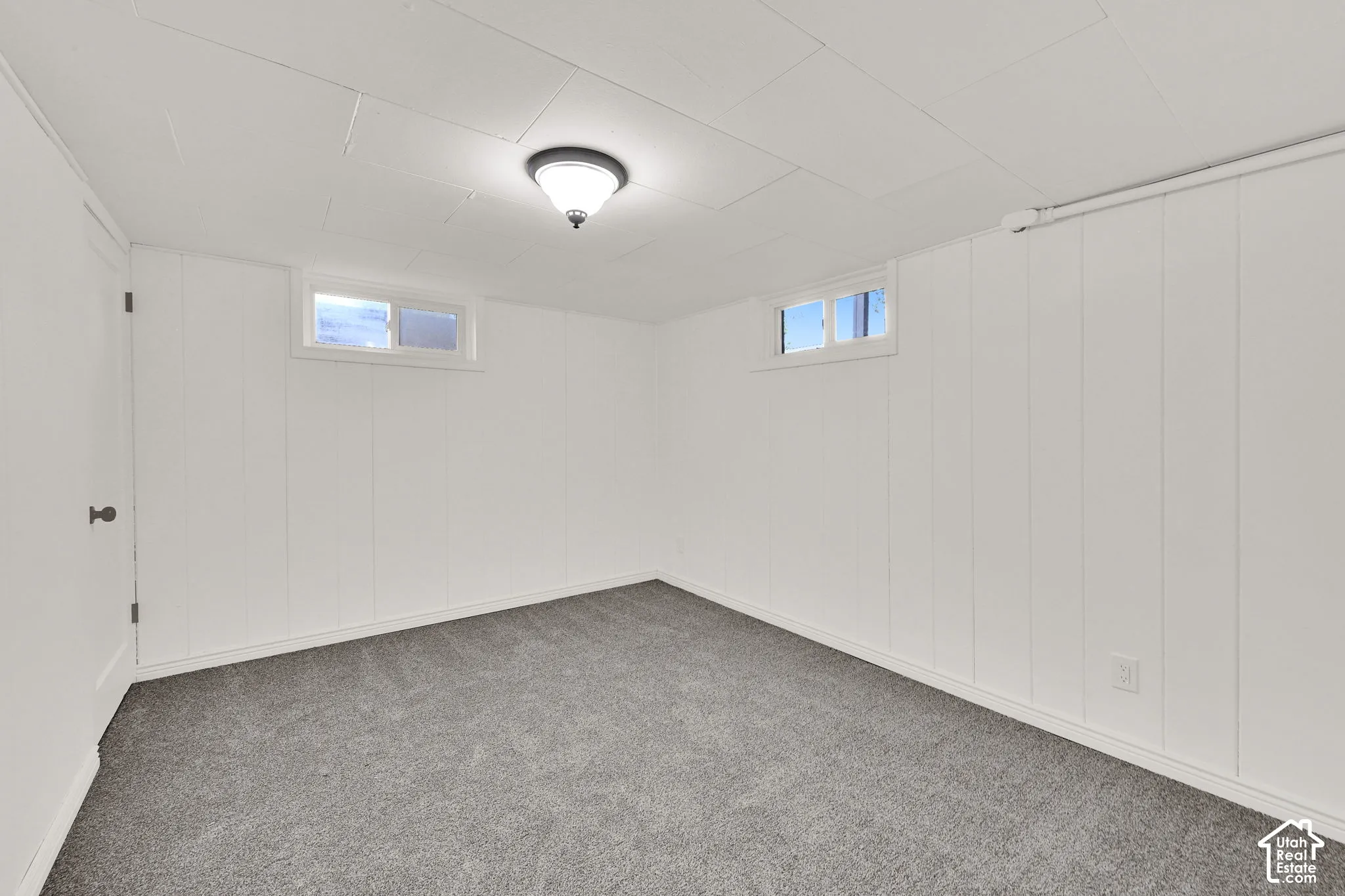 Interior space featuring white vertical wall paneling, neutral carpeting, a tiled ceiling, two rectangular windows, and a flush-mount overhead light fixture