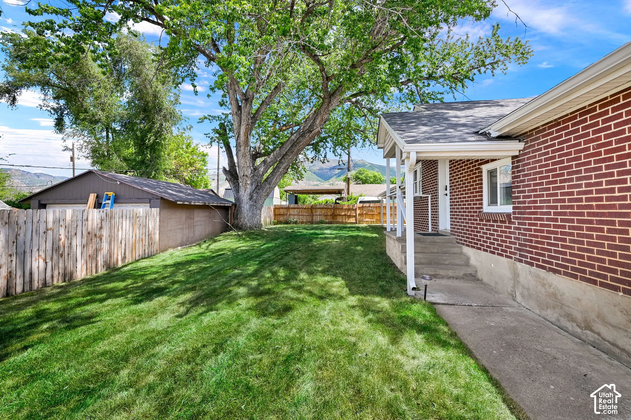 Exterior yard space featuring a brick facade, concrete walkway, mature tree, and wood-panel fencing