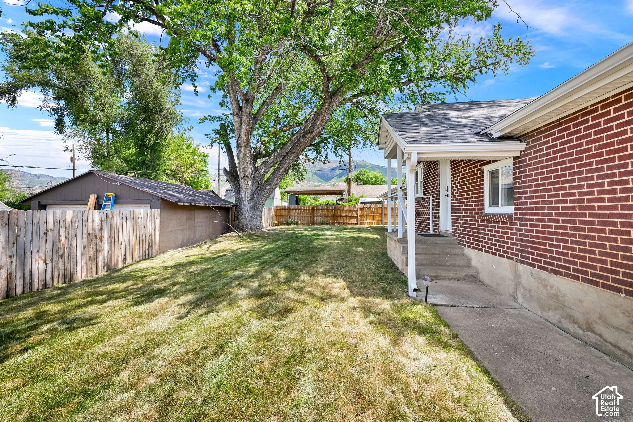 Exterior space featuring brick siding, a concrete walkway, wood fencing, and a mature tree