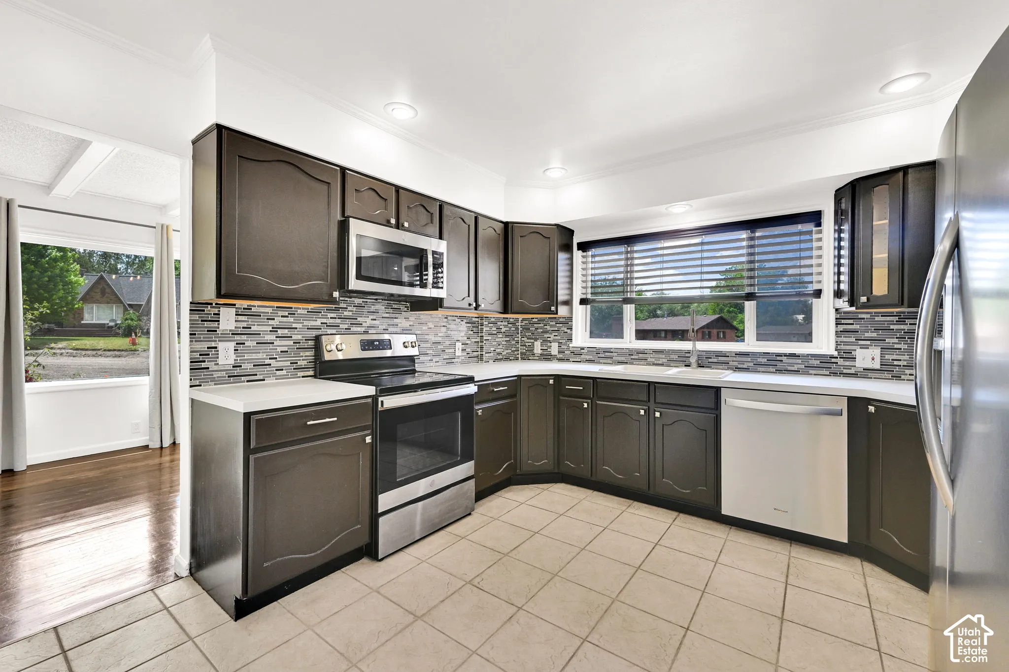 Kitchen featuring dark-toned cabinetry, stainless steel appliances, multi-textured decorative tile backsplash, light-toned solid surface countertops, and neutral tile flooring