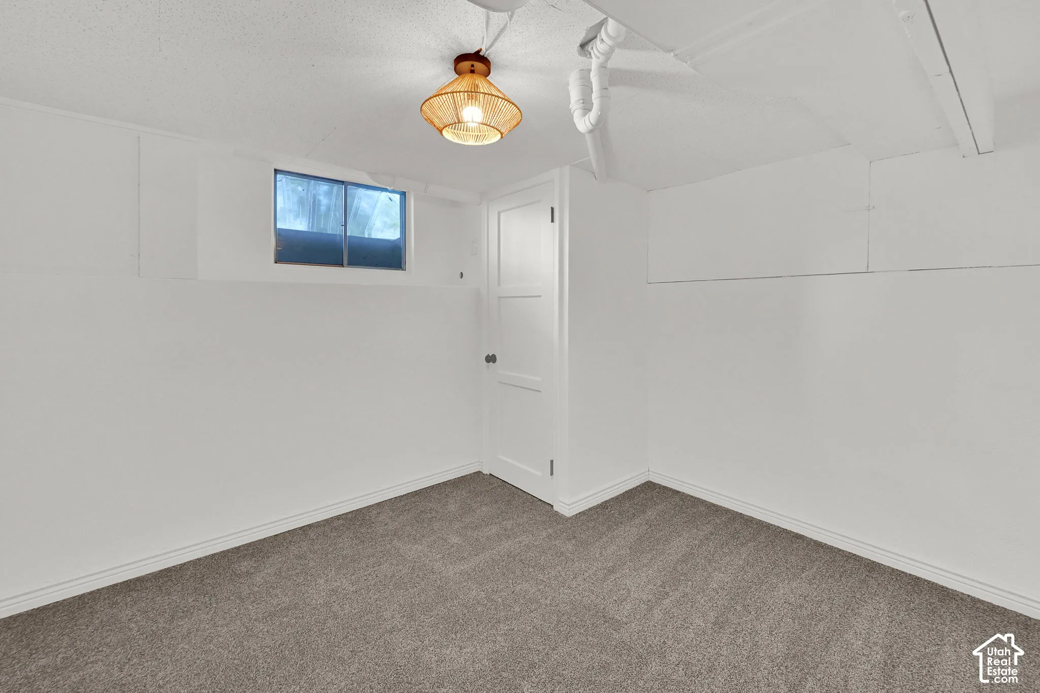 Neutral-toned carpeting, white-painted walls, a single-panel interior door, a rectangular sliding window, and a woven basket-style ceiling light fixture