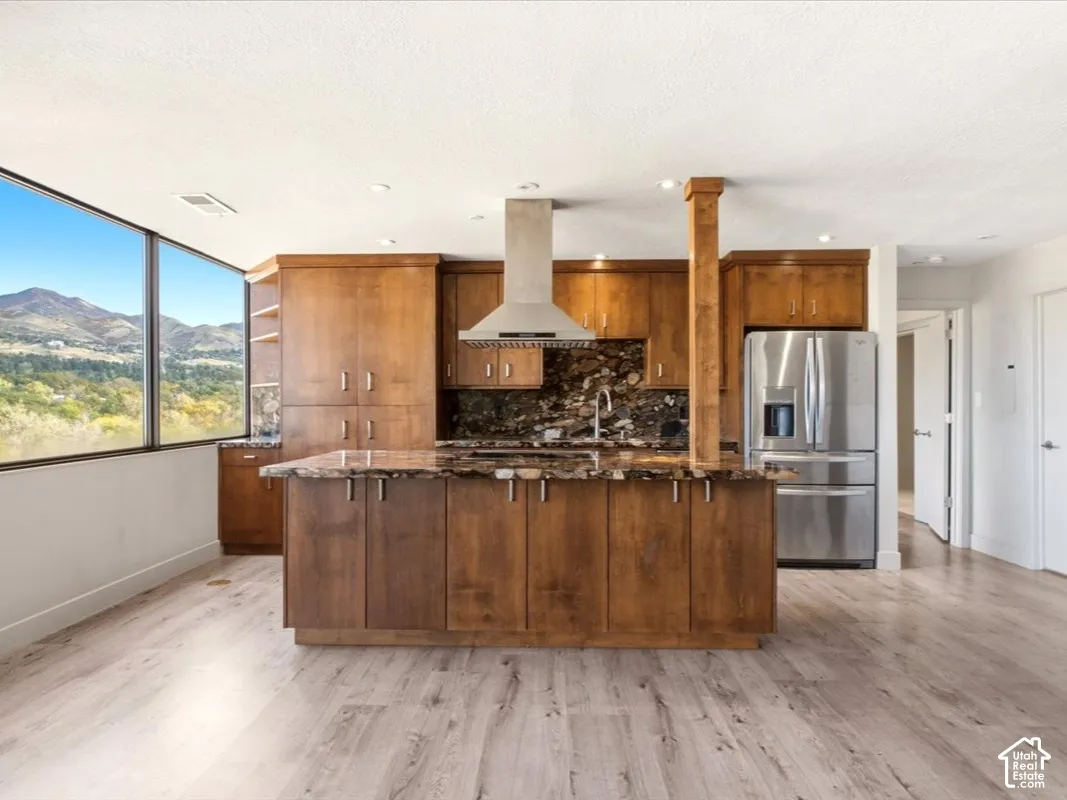 Kitchen featuring wood cabinetry, a central island with a stone-finish countertop, stainless steel appliances, a stainless steel range hood, and floor-to-ceiling windows with mountain scenery