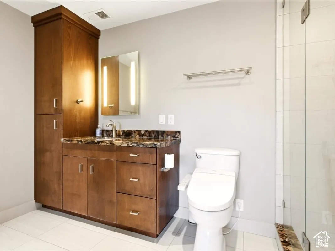Bathroom featuring wood cabinetry with integrated tall storage, stone-patterned countertop, under-mount sink, wall-mounted light fixtures, and a glass-enclosed shower