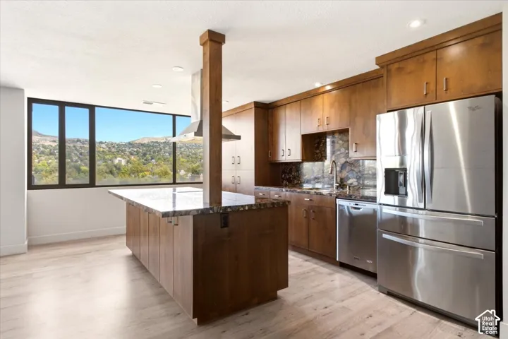 Kitchen featuring custom wood cabinetry, central island with stone-surface countertop, stainless steel appliances, recessed ceiling lighting, and large windows with hillside scenery