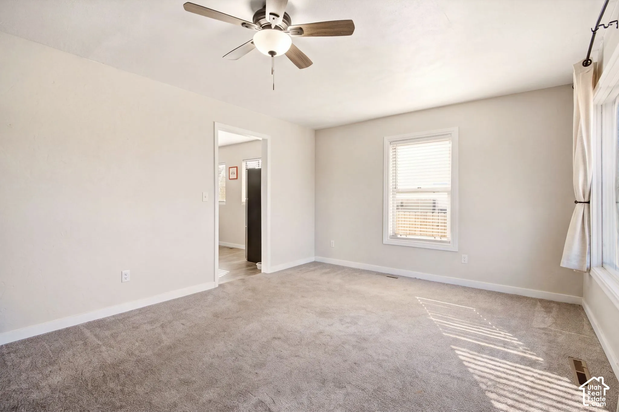 Neutral-toned interior space featuring wall-to-wall carpeting, a multi-blade ceiling fan with integrated lighting, and dual windows