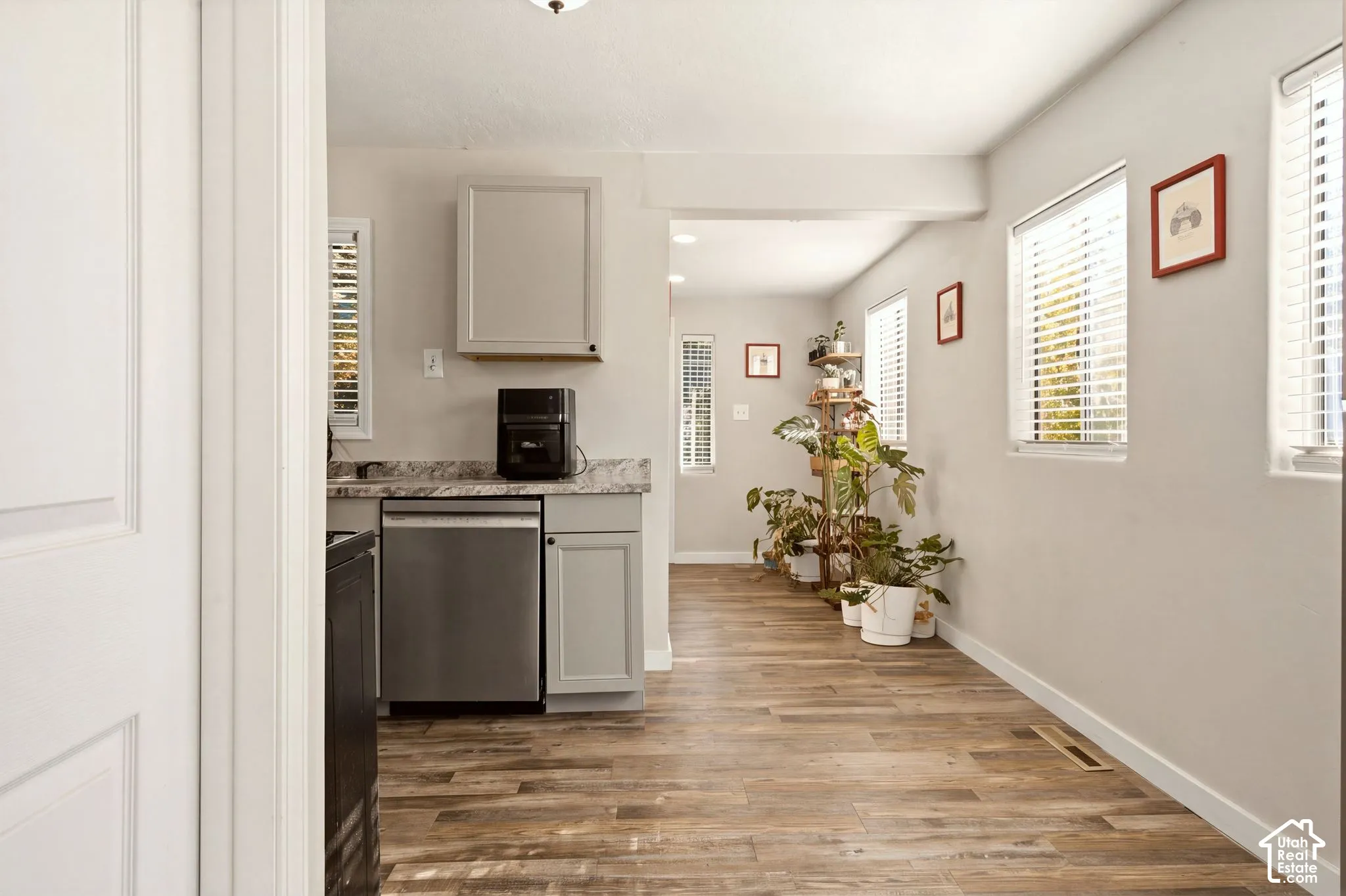Kitchen featuring wood-finish flooring, stainless steel dishwasher, stone-patterned countertops, shaker-style cabinetry, and multiple windows with horizontal blinds