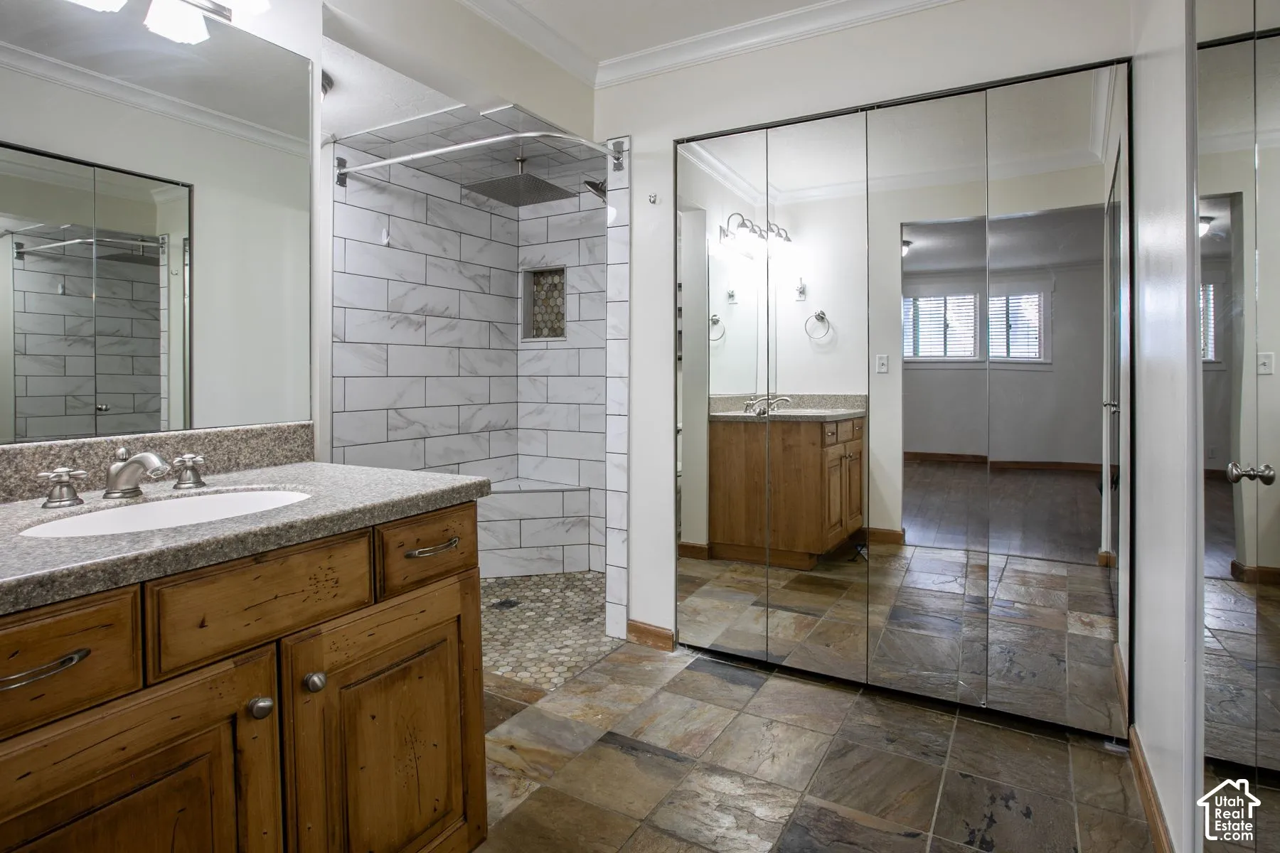 Bathroom featuring a stone-tile floor, wood-finish vanity with a stone countertop, walk-in shower with white subway-style tile walls and a pebble-stone floor, and floor-to-ceiling mirrored closet doors