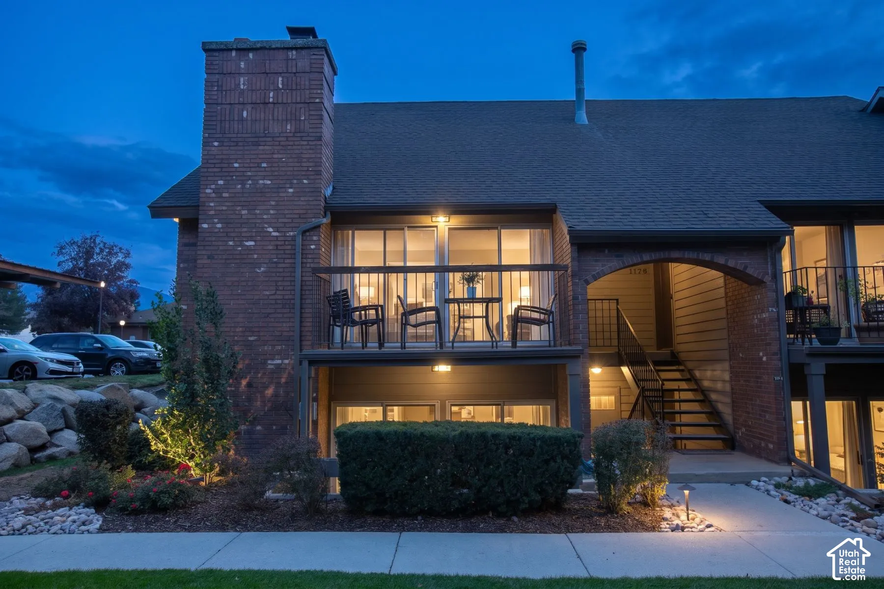 Brick exterior featuring a multi-story layout, a prominent chimney, an elevated balcony with railing, and a stone-accented landscape