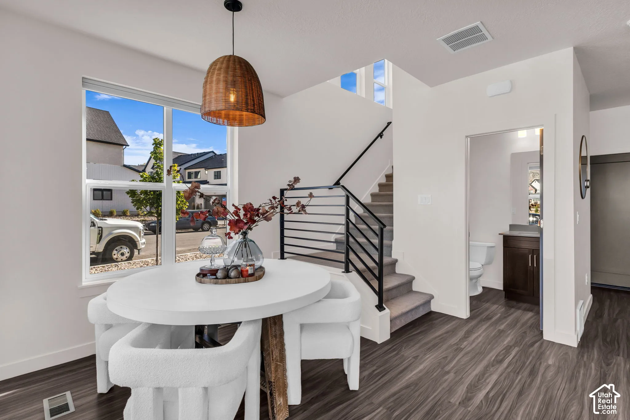 Wood-finish flooring, staircase with metal horizontal railing, large windows, woven pendant light fixture, and bathroom with dark wood-finish cabinetry