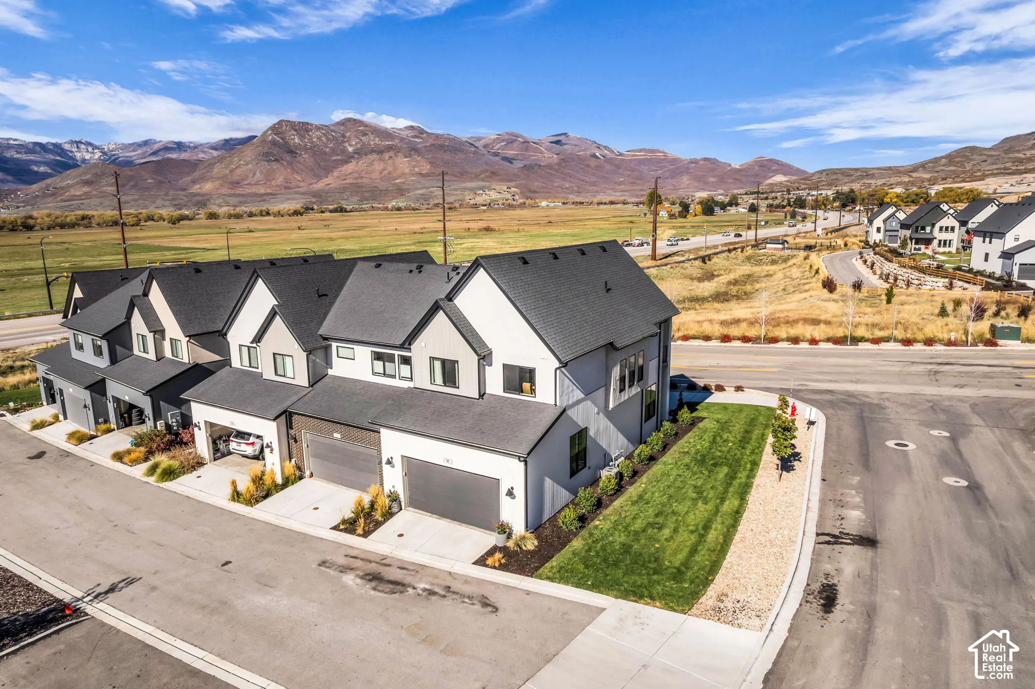 Multi-story townhouse exterior featuring white siding with dark trim, asphalt shingle roofing, attached garages, and a landscaped corner lot with mountain scenery