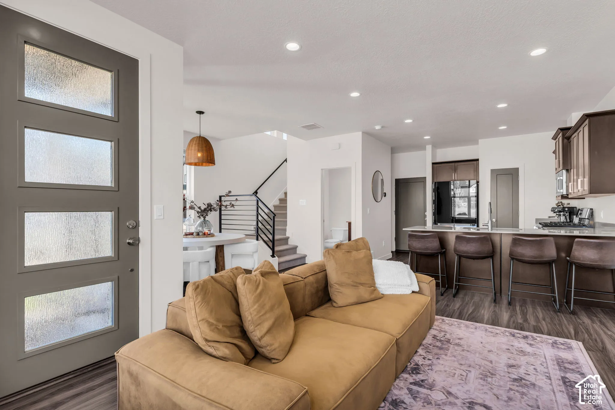 Modern entry featuring a gray door with four frosted glass panels, wood-finish flooring, recessed lighting, dark cabinetry with breakfast bar seating, and a staircase with metal horizontal railing