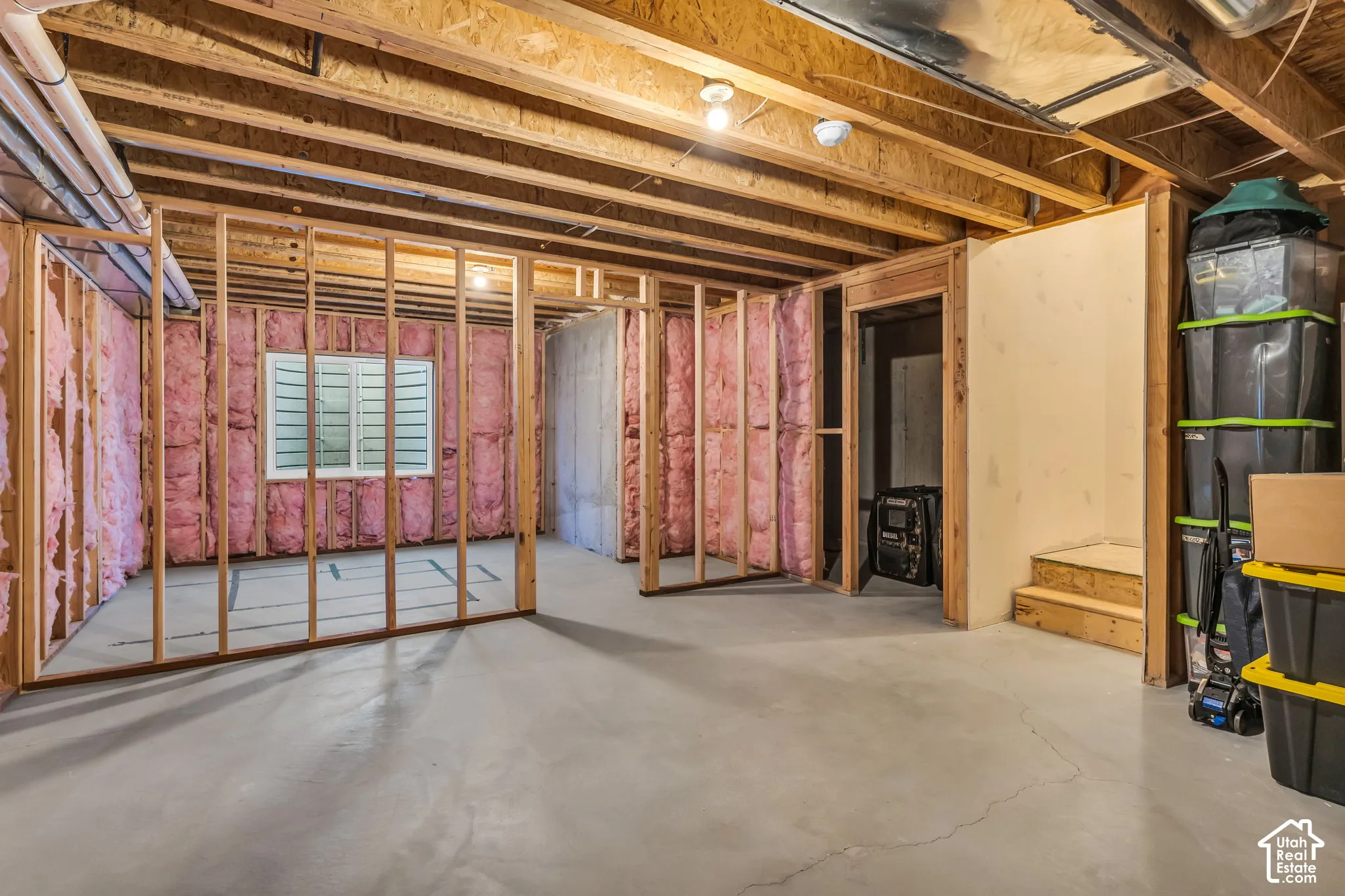 Unfinished basement featuring exposed wood joists, pink batt insulation, concrete flooring, and an egress window