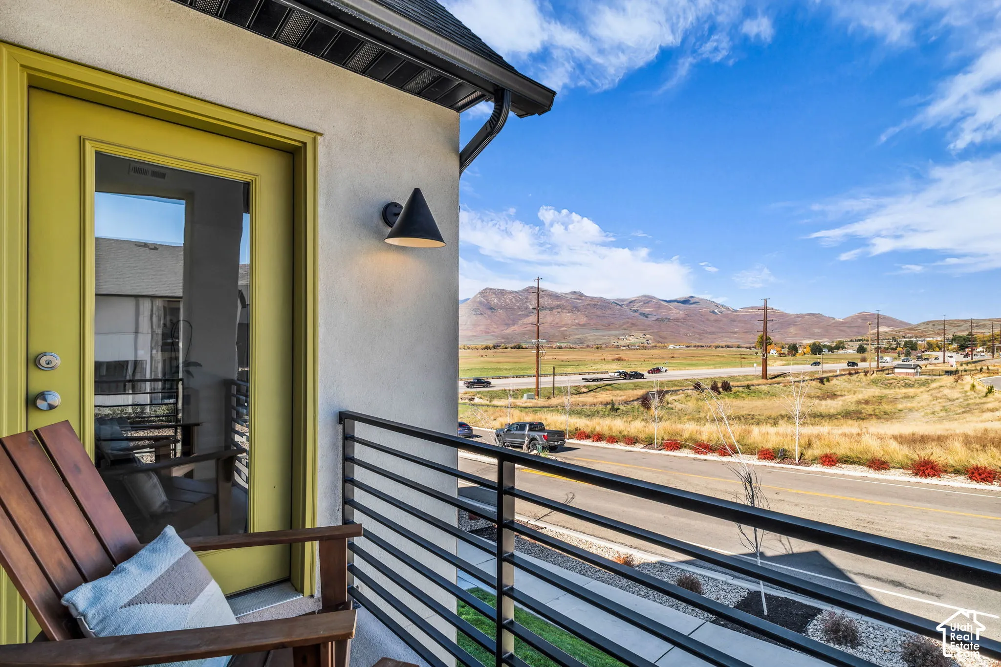 Balcony featuring a horizontal metal railing, exterior wall-mounted conical lighting, and a painted door, set against a mountain backdrop
