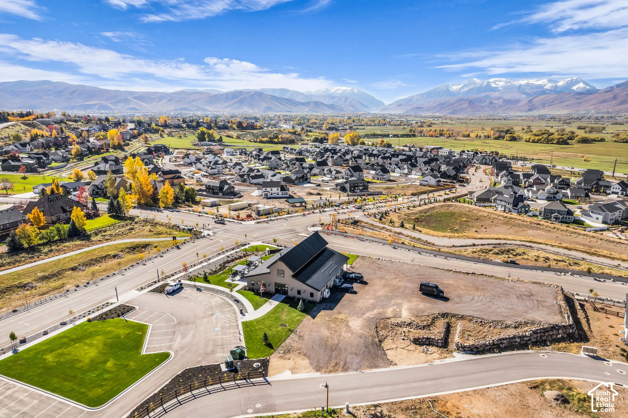 Aerial perspective featuring a clubhouse with a metal roof, paved parking area, manicured lawn, stone retaining wall, and mountain backdrop